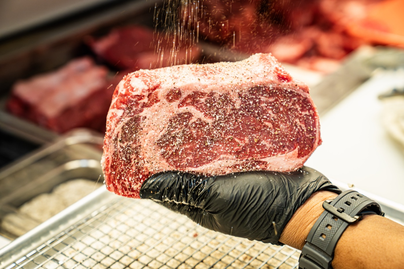 A butcher at Little Alley Steak Roswell hand-seasoning a freshly cut USDA Prime bone-in steak, cracked pepper and salt falling across the marbled surface